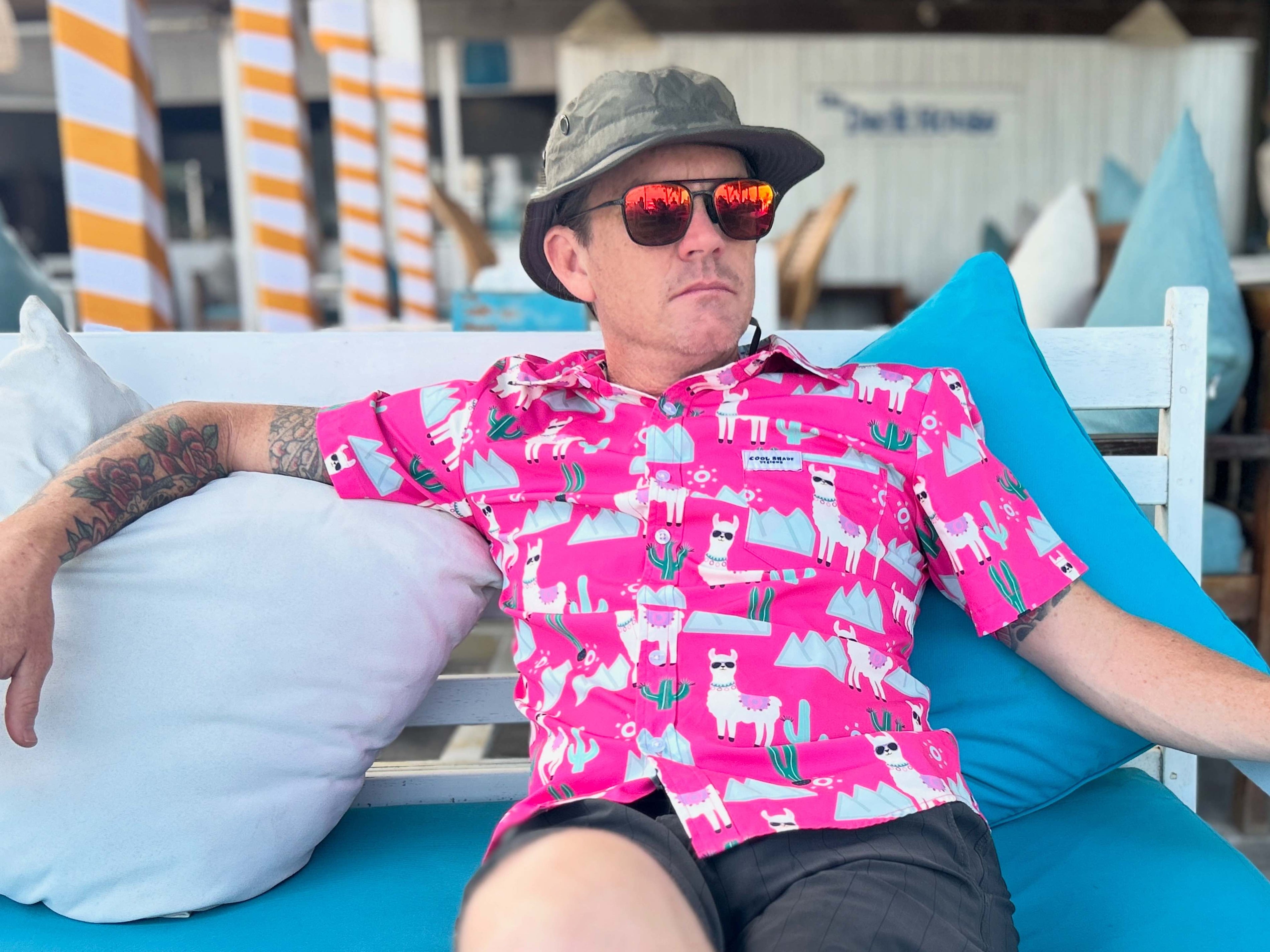 Man wearing a bright pink shirt with llama pattern, sunglasses, and a cap sitting on a blue chair. A nod To Phish's song Llama.