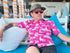Man wearing a bright pink shirt with llama pattern, sunglasses, and a cap sitting on a blue chair. A nod To Phish's song Llama.