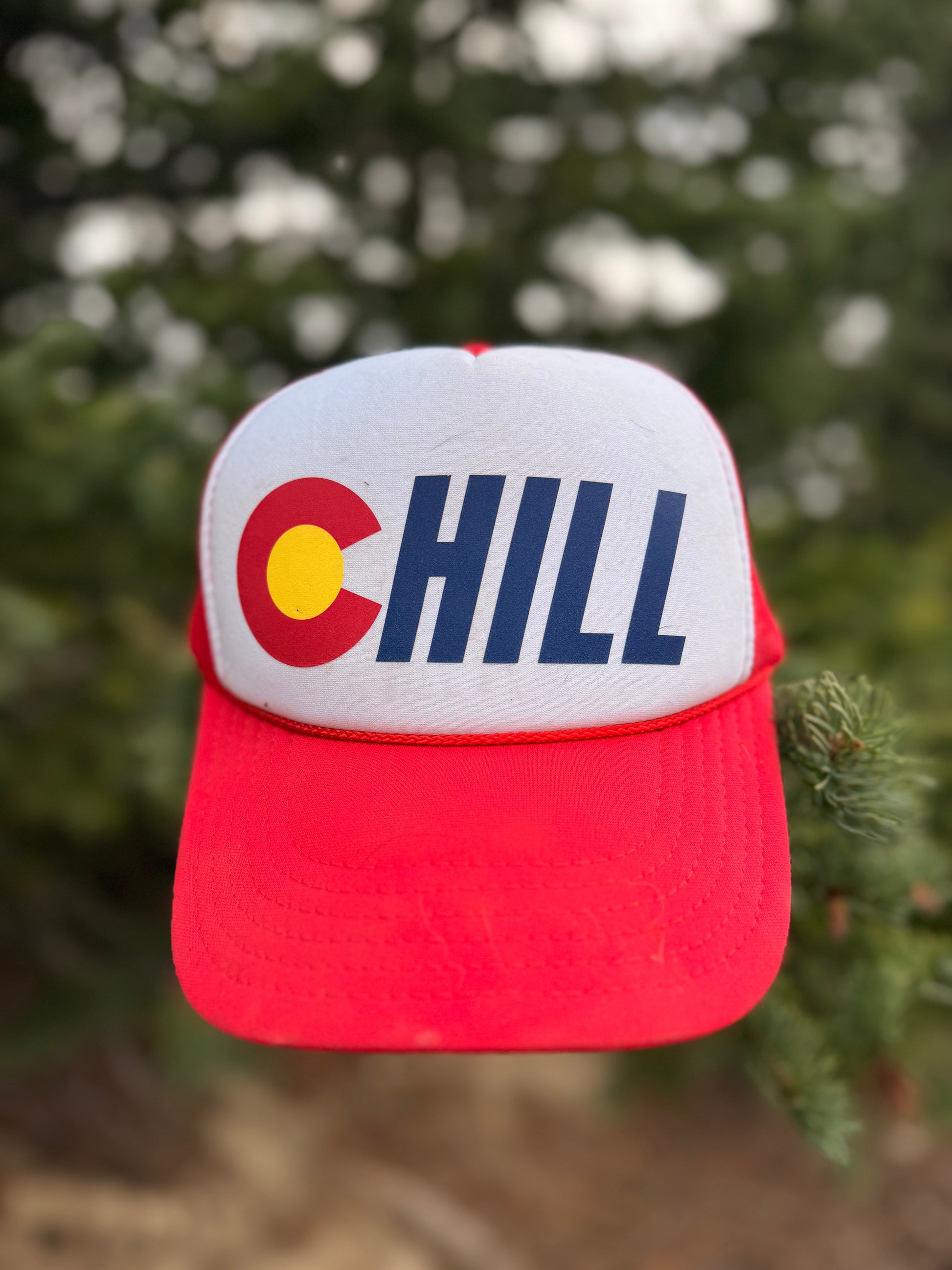A red and white trucker-style cap with a Colorado 'C' and the word 'CHILL' on the front, photographed outdoors against a blurred green background.
