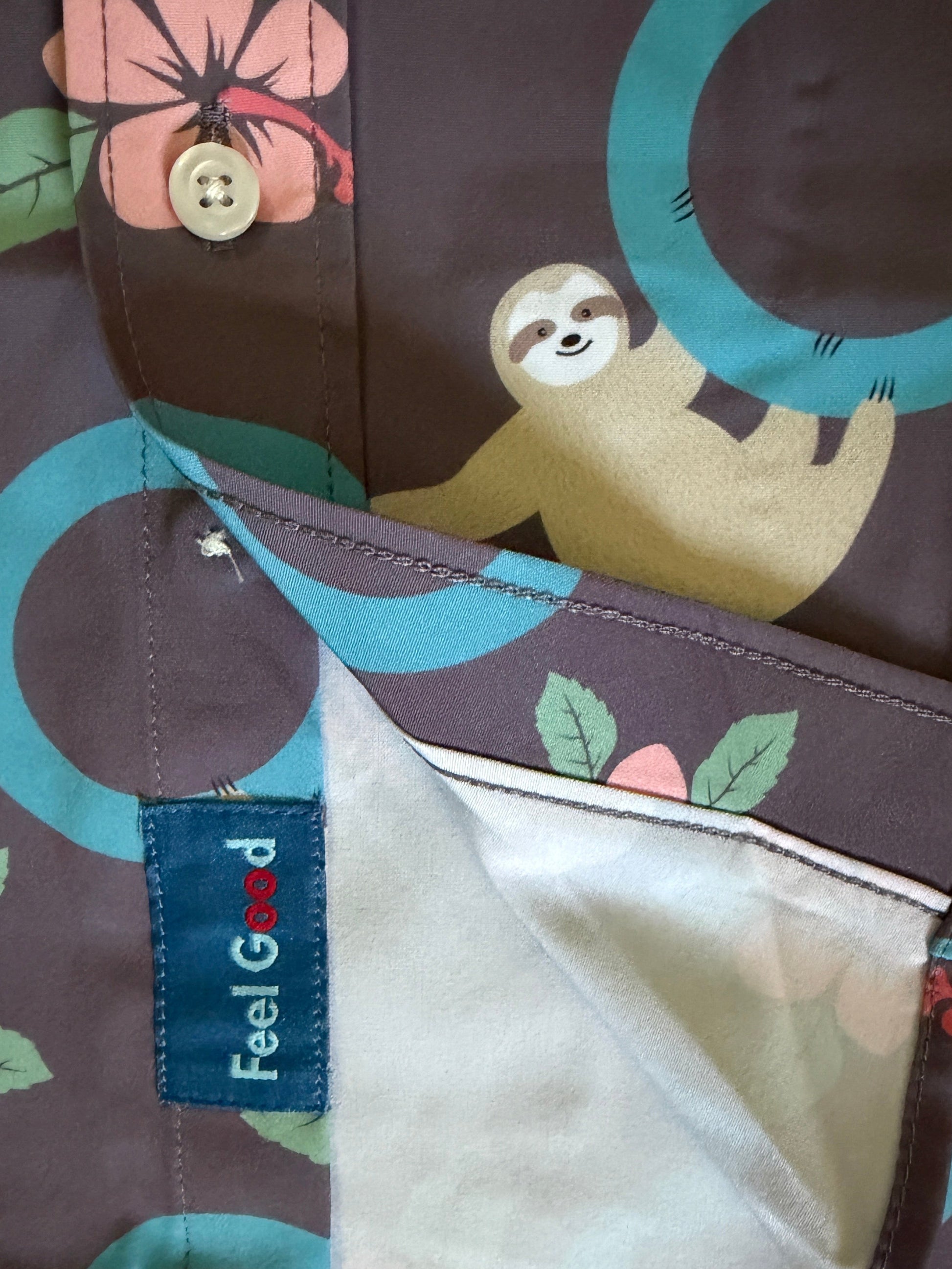 Close-up of a patterned button-up shirt laid flat r. The fabric is dark brown with a colorful print of a sloth, teal circles, pink flowers, and green leaves.